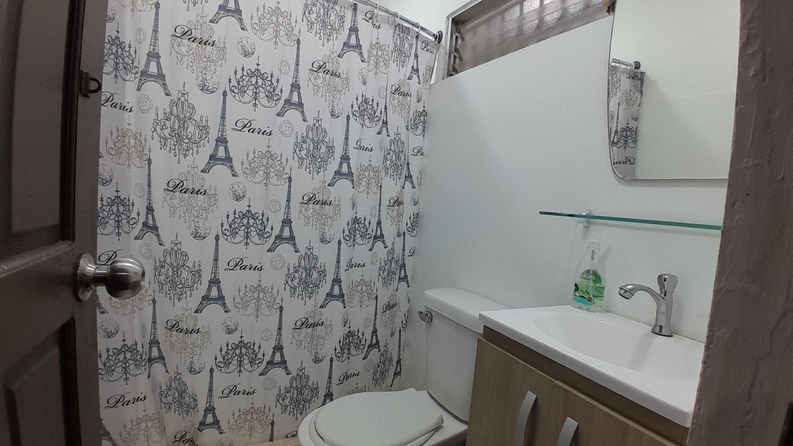 A bathroom with a shower curtain featuring the Eiffel Tower and chandeliers, a toilet, a sink with a glass shelf, and a mirror.