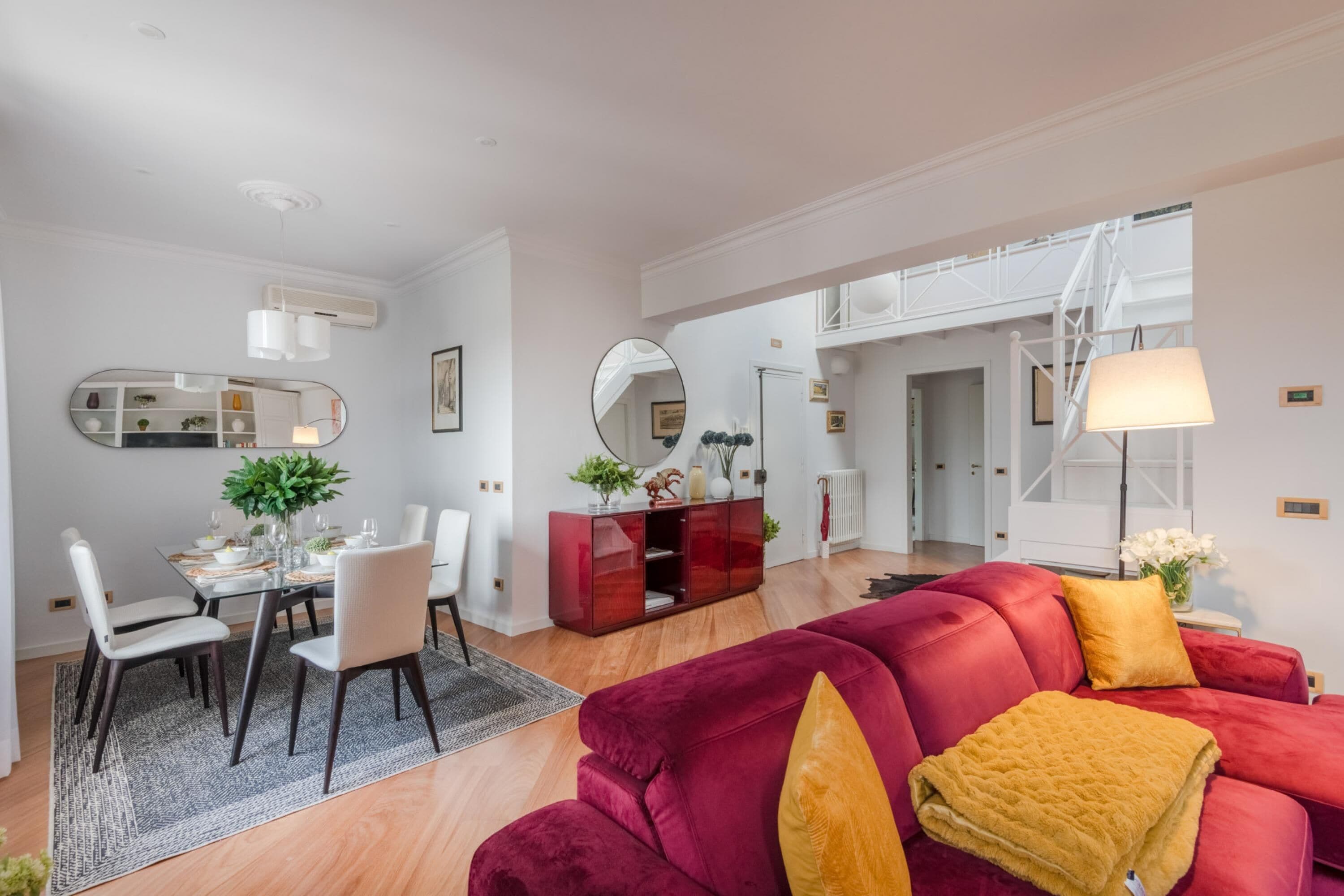 Una sala de estar moderna con un sofá rojo, un comedor con una mesa de vidrio y un pasillo que conduce a una escalera.