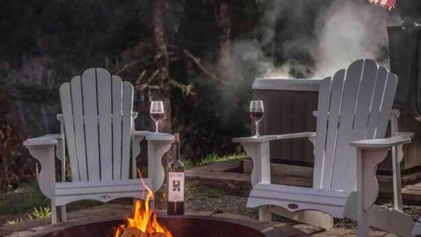 Zwei Adirondack-Stühle mit einem Feuerkorb dazwischen, eine Weinflasche und ein Glas auf jedem Stuhl.