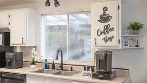 Eine moderne Küche mit weißen Schränken, einer Wanddekoration zum Thema Kaffee, einer Kaffeemaschine und einem Fenster mit Jalousien.