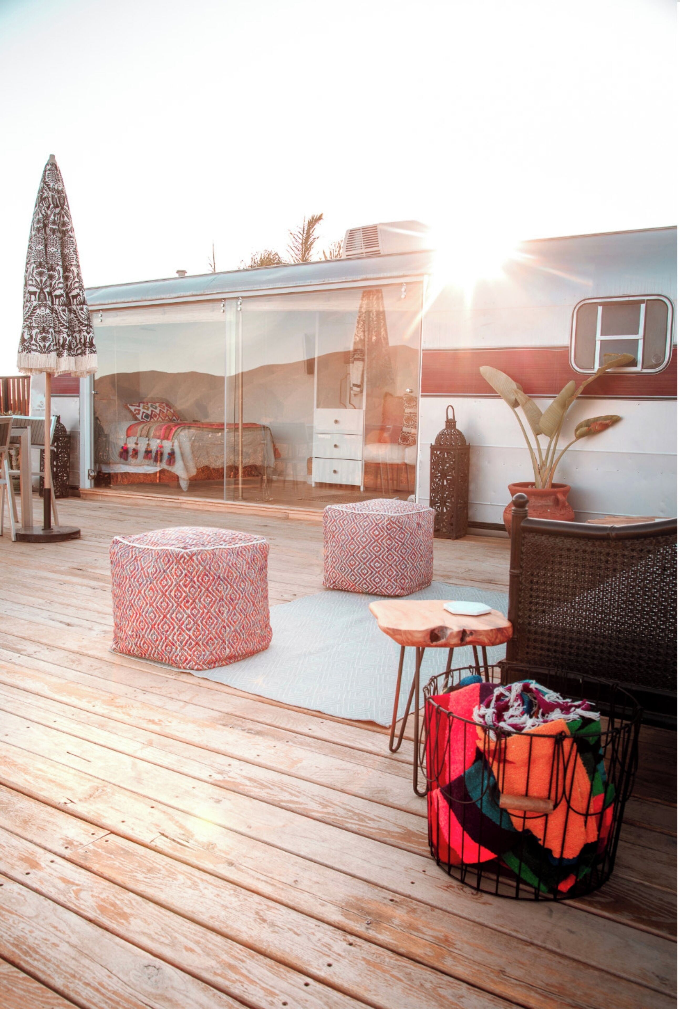 A cozy outdoor deck with wooden flooring, two patterned ottomans, a wicker chair, a small table, and a potted plant.