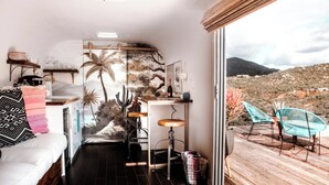 A compact room with a bed, a small bar area, and a view of a desert landscape through a large window. - Arre Lulu Baja (Valle de Guadalupe)