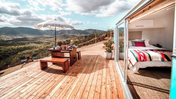 A wooden deck with a table and chairs, a potted plant, and a bed with a colorful blanket. - Arre Lulu Baja (Valle de Guadalupe)