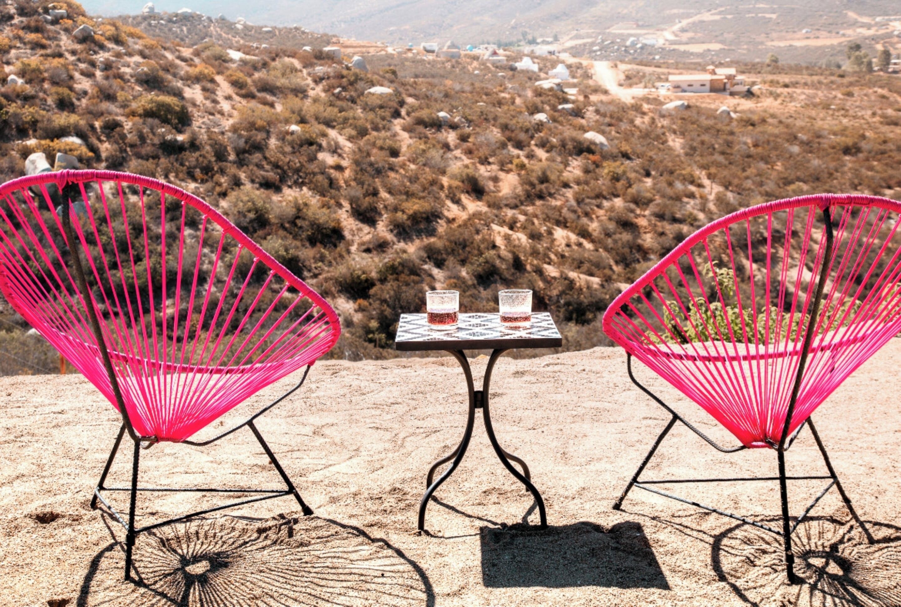Two pink wire chairs and a small table with two glasses on a sandy surface, overlooking a hilly landscape.