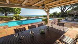 Área da piscina com mesa de madeira, cadeiras e vista para o mar.