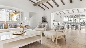 Uma sala de estar espaçosa com um sofá branco, uma mesa de centro e uma poltrona. É possível ver uma área de cozinha através de um arco aberto.