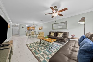 Un salon avec un canapé en cuir marron, une table basse en bois, un tapis à motifs, un ventilateur de plafond et, au fond, une cuisine.