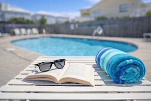 Cenário à beira da piscina com uma espreguiçadeira, um livro aberto e uma toalha enrolada.