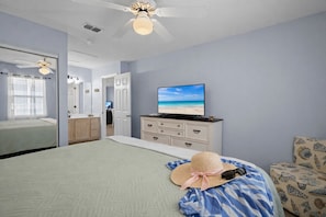 Quarto com cama, televisão em uma cômoda, ventilador de teto e vista para a praia pela janela.