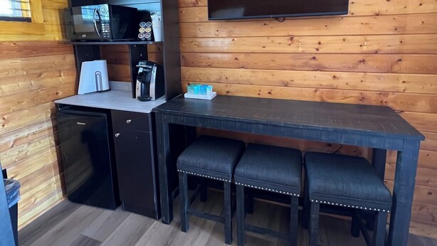 A compact kitchen with a microwave, coffee maker, and a small refrigerator. There is a wooden dining table with three chairs and a flat-screen TV mounted on the wall.