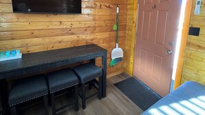 A room with a wooden wall, a flat-screen TV mounted above a dark wooden table with benches, a broom, and a door with a handle.