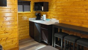 A compact kitchen with wooden paneling, a microwave, a coffee maker, and a small sink. There is a bar area with stools and a ceiling-mounted air conditioner. - Sandstone Cabins at the Red Desert RV Park (Moab)