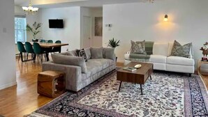 A spacious living room with a grey sofa, a white sofa, a wooden coffee table, and a patterned rug. There is a dining area with a table and chairs in the background.