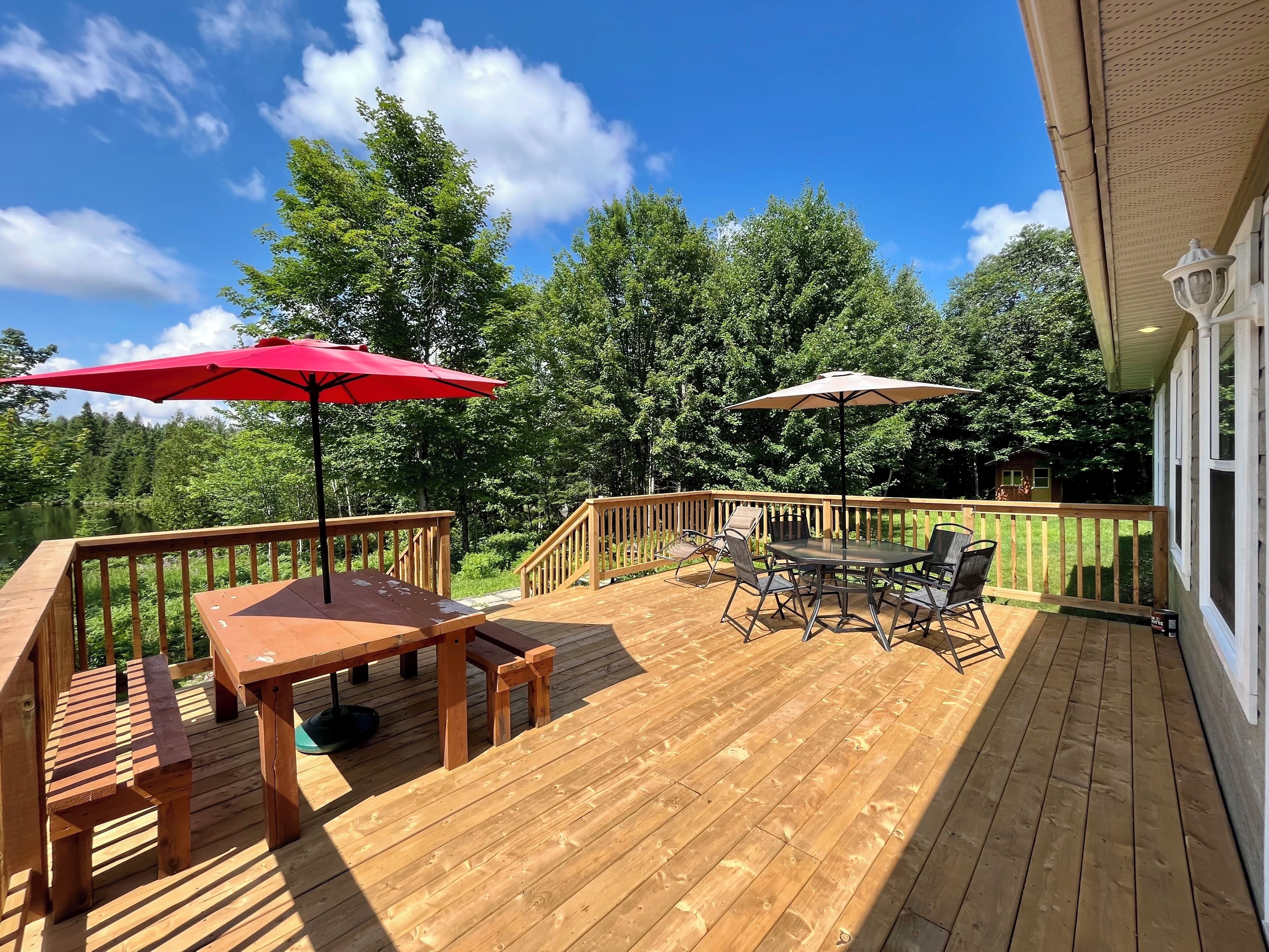 A wooden deck with a table and chairs, two umbrellas, and a view of a lake and forest.
