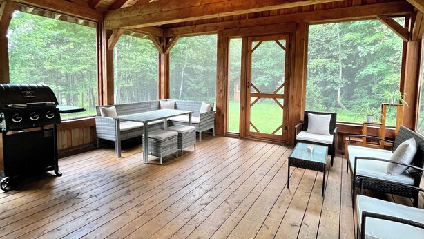 A wooden deck with outdoor furniture, a grill, and large windows offering a view of the forest.