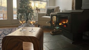 A cozy living room with a fireplace, two glasses of wine on a wooden table, and a Christmas tree in the background.