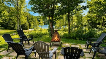 Un espace extérieur aménagé avec des chaises Adirondack et un foyer, entouré d’une végétation luxuriante et offrant une vue sur un lac.