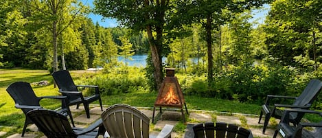 Ein Sitzbereich im Freien mit Adirondack-Stühlen und einem Feuerkorb, umgeben von üppiger Vegetation und Blick auf einen See.