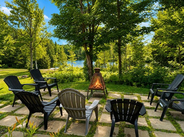 An outdoor seating area with Adirondack chairs and a fire pit, surrounded by lush greenery and a lake view.