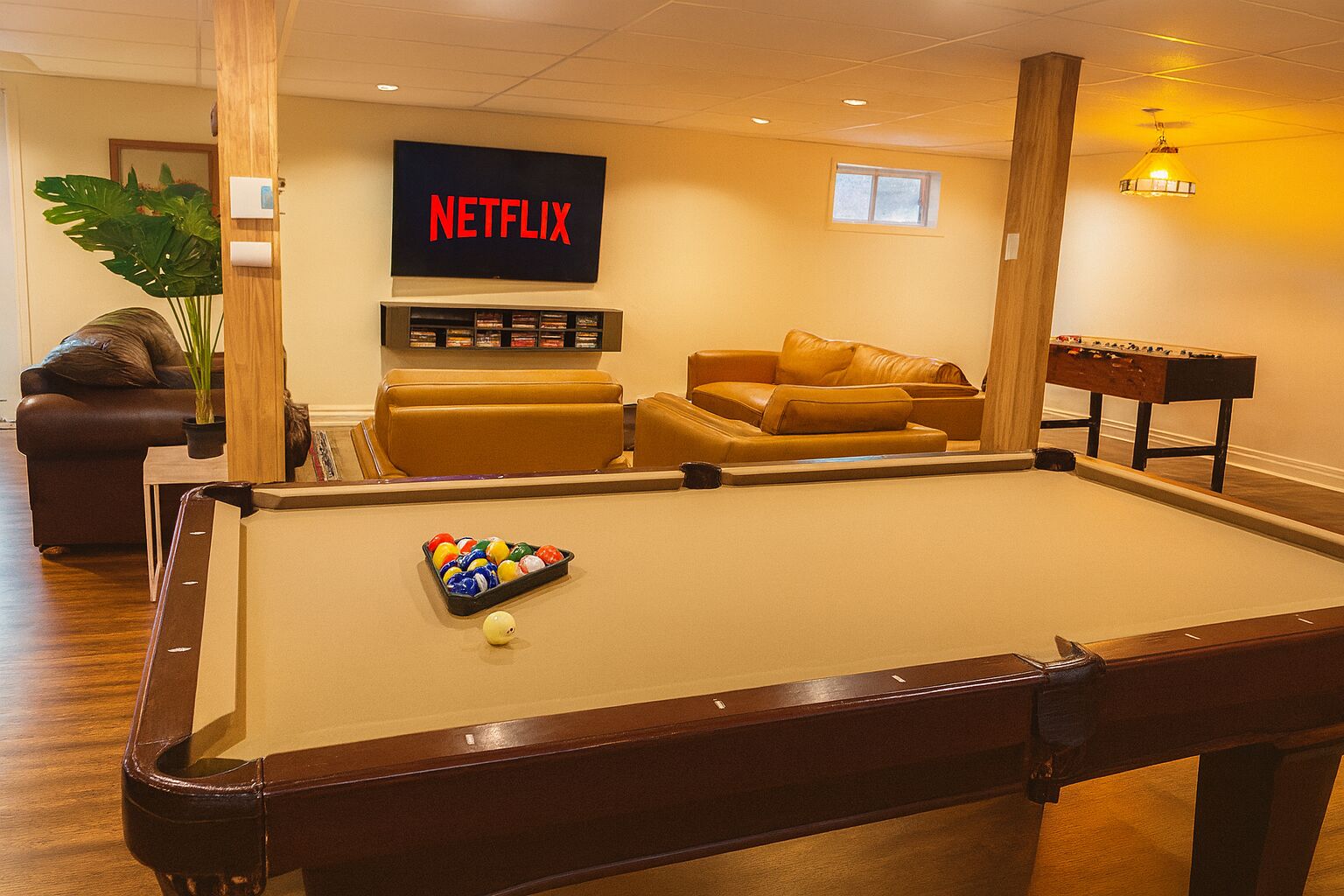Uma mesa de bilhar com bola branca e bolas de bilhar, um sofá, uma mesa de pebolim, uma televisão exibindo o logotipo da Netflix e um vaso de plantas.