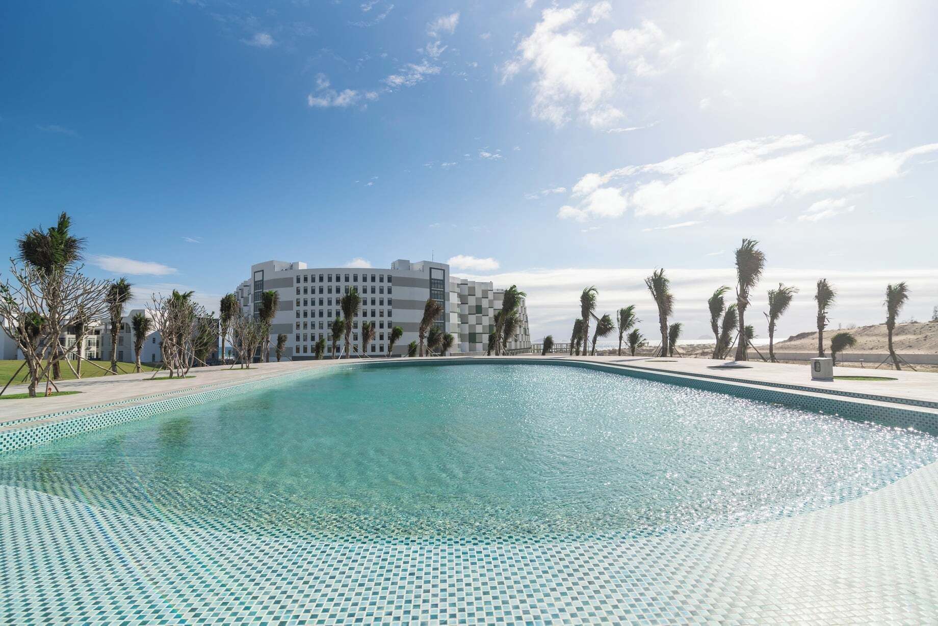Ein Swimmingpool mit gefliestem Rand, umgeben von Palmen und einem Gebäude mit moderner Architektur im Hintergrund.