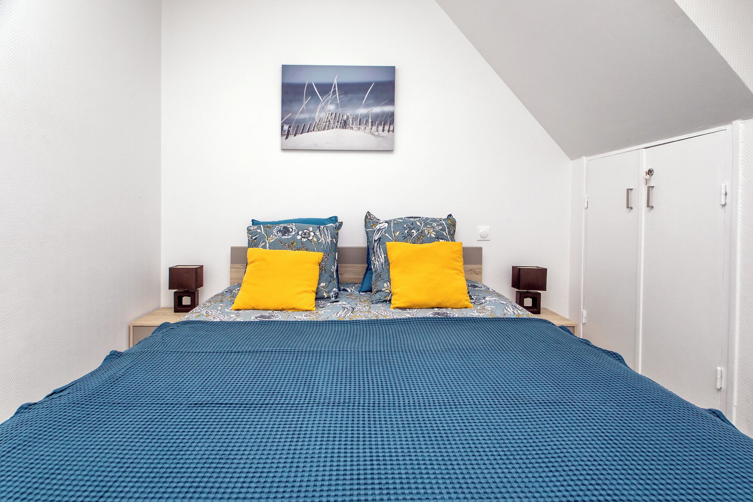 A bedroom with a large bed, two nightstands, a painting on the wall, and a wardrobe.