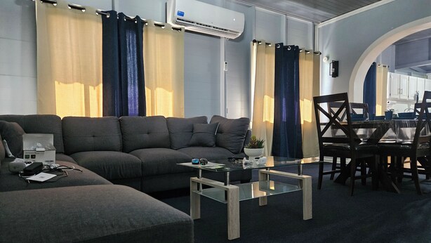 Een moderne woonkamer met een grijze bank, een glazen salontafel, en een eethoek met houten stoelen en een tafel.