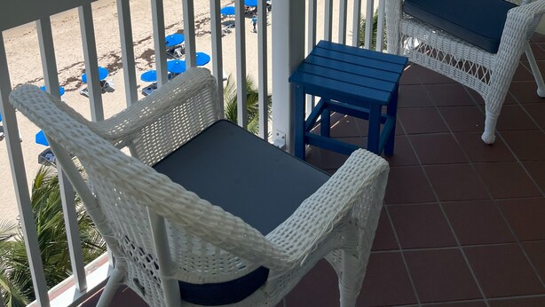 Een balkon met witte rieten stoelen en een kleine tafel, met uitzicht op een strand met blauwe parasols en een helder uitzicht over de oceaan.