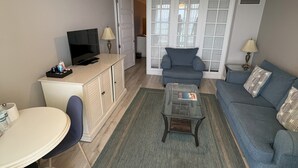 Een woonkamer met een bank, salontafel en televisie. Er is een eethoek met een tafel en stoelen. De kamer heeft een plafondventilator en een raam met jaloezieën.