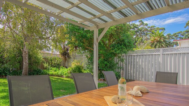 Une table en bois placée à l’extérieur, avec des chaises, une bouteille et un verre, le tout installé sous une pergola offrant une vue sur un jardin et une clôture.