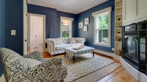 Un salon avec un canapé blanc, des fauteuils à motifs, un pouf à carreaux, une lampe sur pied et une cheminée.