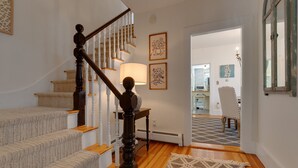 Un escalier avec une rampe en bois et des marches recouvertes d’une moquette beige, menant à une pièce où se trouvent un tapis à motifs et une table avec une lampe.