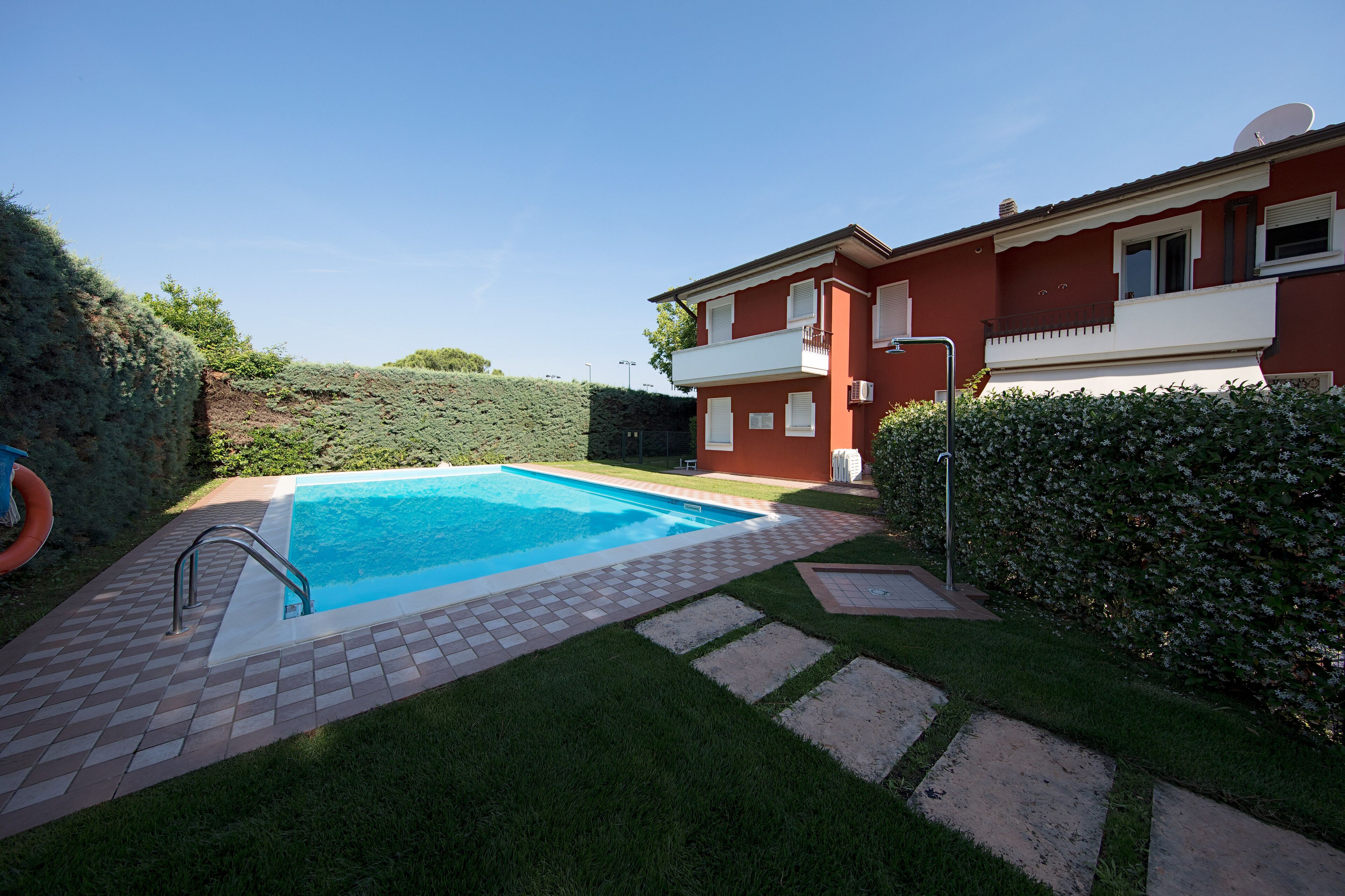 Une piscine avec une terrasse carrelée, un bâtiment rouge doté d’un balcon et une haie.