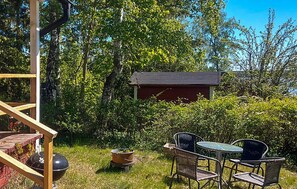 Une terrasse en bois avec un barbecue, un petit abri de jardin et une table avec des chaises, le tout situé dans un jardin luxuriant.