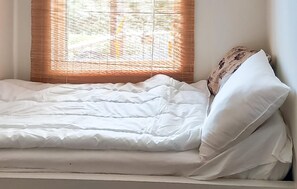 Uma cama com roupa de cama branca, uma janela com persianas e um travesseiro floral.