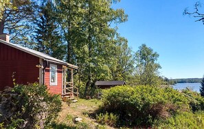 A red cabin with a porch, surrounded by dense greenery and a lake. - 1 bedroom cozy home in Eskilstuna (Eskilstuna)
