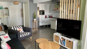 Un salon moderne avec un canapé, deux petites tables, un téléviseur à écran plat et une cuisine en arrière-plan.