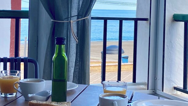 Une table avec des aliments pour le petit-déjeuner, une bouteille verte et une vue sur la plage et l’océan.