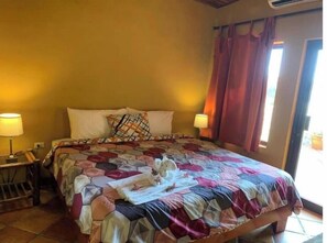 A hotel room with a large bed, two bedside lamps, a window with curtains, and a small table with a lamp. - Infinity Lounge Party's House (Garabito)
