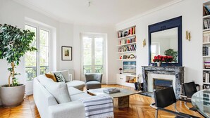 A spacious living room with a large sofa, a coffee table, and a fireplace. There are bookshelves filled with books and a large mirror above the fireplace.