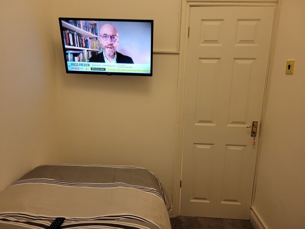 A room with a bed, a television mounted on the wall displaying a news program, and a closed door. - Timann Place (Manchester)