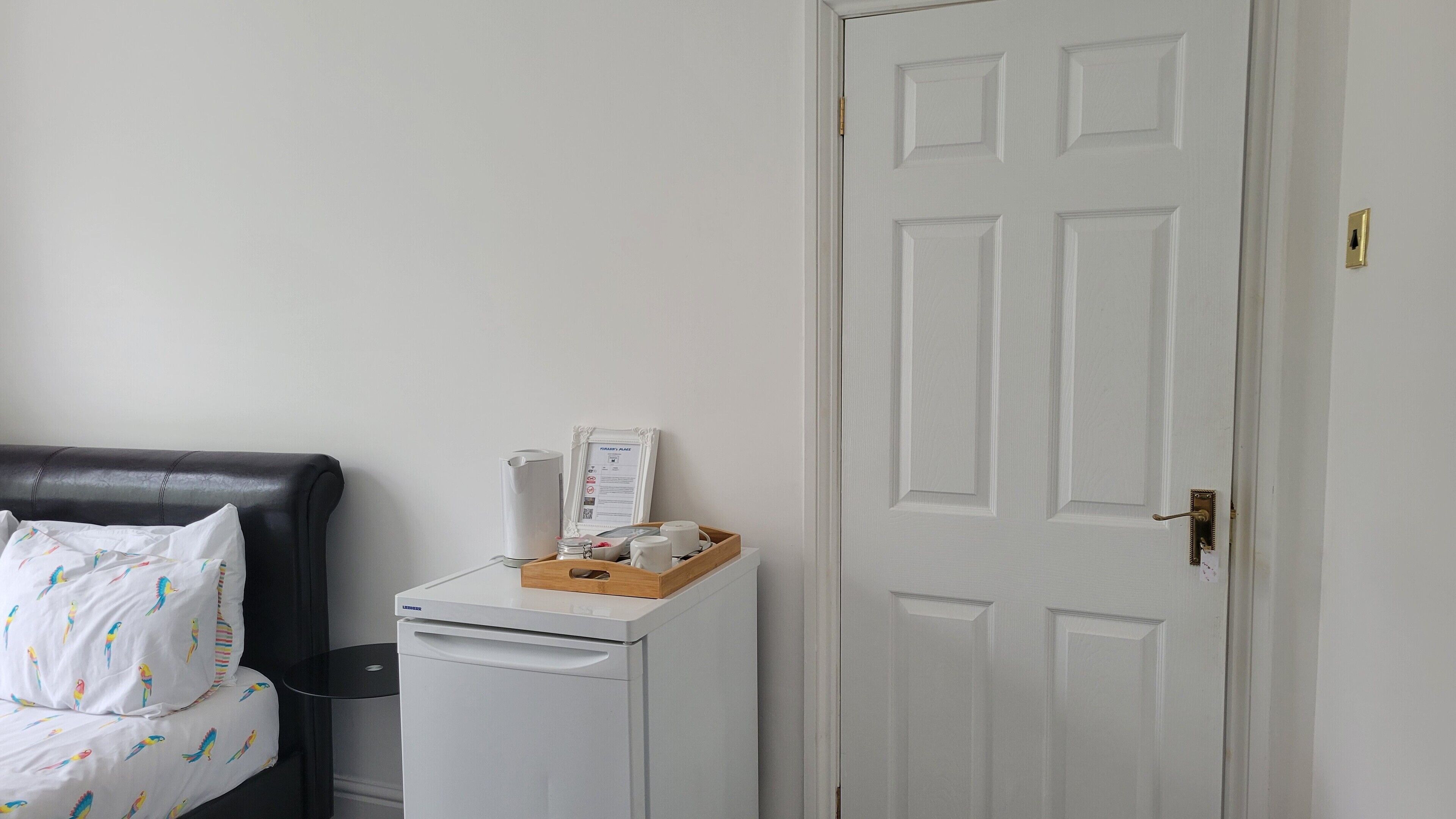 A bedroom with a bed, a small white bedside table with a lamp, a framed certificate, and a door.