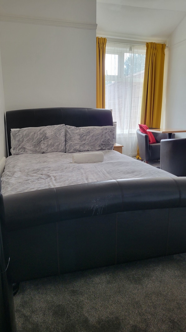 Un lit bien fait, avec une tête de lit noire et du linge de lit blanc, une petite table de chevet avec une lampe et une fenêtre ornée de rideaux jaunes.