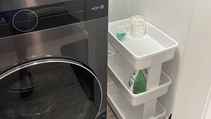 Un espace buanderie compact comprenant un lave-linge et un sèche-linge à chargement frontal, une petite étagère blanche avec des récipients et une décoration murale à motifs.