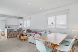 A modern kitchen with white cabinets, a dining area with a wooden table and chairs, and a grey sofa with cushions.