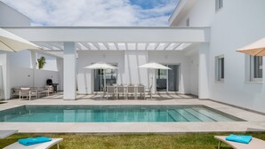 Un área de piscina exterior moderna con una zona de estar cubierta, una parrilla y un cielo despejado.
