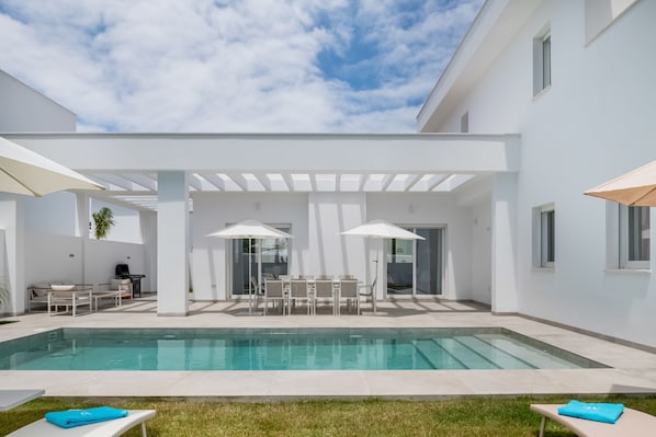 A modern outdoor pool area with a covered seating area, a grill, and a clear sky.