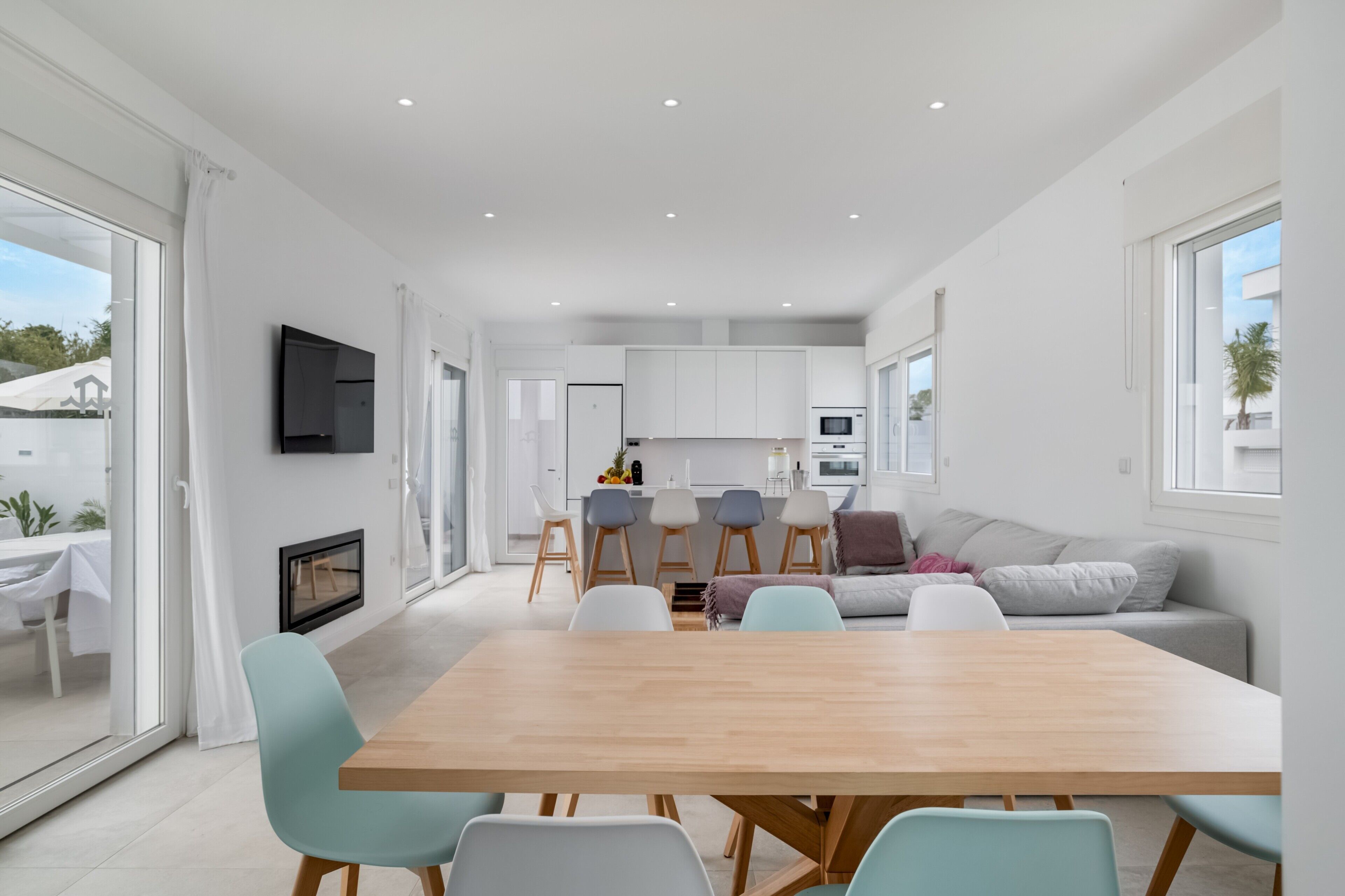 A modern kitchen with white cabinets, a dining area with a wooden table and chairs, and a living area with a sofa and a TV.
