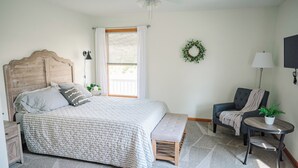 Un dormitorio con una cama de madera, un banco, una silla, una mesita con una planta, una lámpara de pared y una ventana con cortinas.