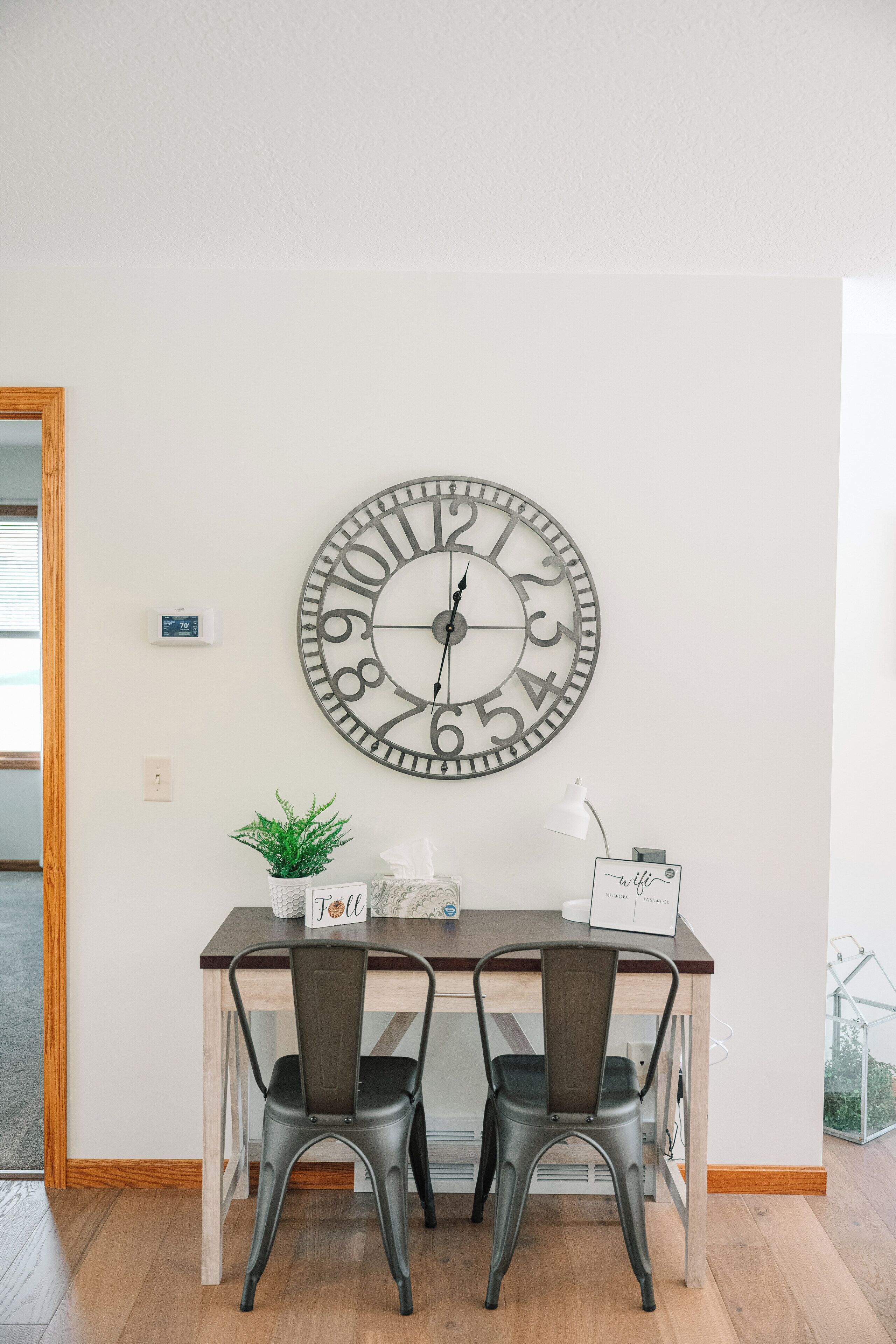 A dining area with a wooden table, two chairs, a wall clock, a potted plant, and a Wi-Fi sign.